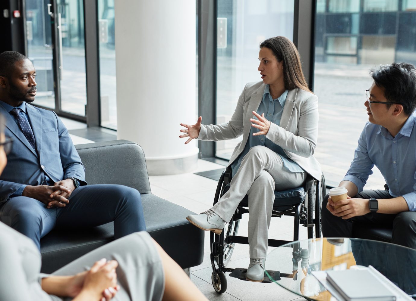 Groupe divers de personnes d'affaires se réunissant dans le hall d'accueil d'un bureau, avec un focus sur une jeune femme en fauteuil roulant partageant des idées / Diverse group of business people meeting in office lobby with focus on young woman in wheelchair sharing ideas